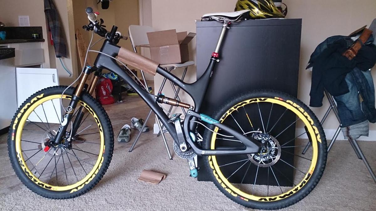Yeti SB66c: A mountain bike with a distinctive wooden section on the frame is leaned against a black piece of furniture. The bike features large yellow tires and is positioned in a room with various items visible in the background, including a backpack, shoes, and some clothing.