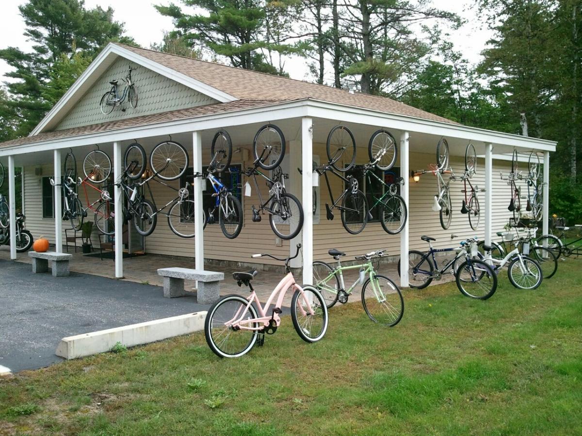 A creative display of various bicycles hanging on the exterior of a building, with additional bikes parked on the ground. The building has a light-colored exterior and a sloped roof, surrounded by grass and tall trees in the background. A small pumpkin is placed on the porch, adding a seasonal touch.