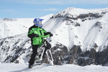 A person dressed in a bright green and black ski outfit, wearing a blue helmet, is standing on a snowy mountain slope. They are riding a snow bike, with snow-covered peaks and a clear blue sky in the background.