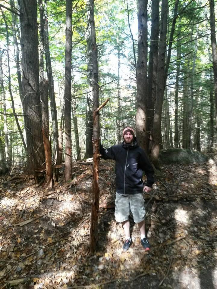 A man standing in a wooded area, leaning against a tree while wearing a black zip-up jacket and shorts. The ground is covered with fallen leaves, and the surrounding trees are tall and green, indicating a sunny day.