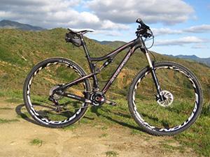 Santa Cruz Tallboy Carbon: A mountain bike positioned on a dirt trail, with green hills and a blue sky in the background. The bike features a sleek, black frame and large, rugged tires designed for off-road terrain.