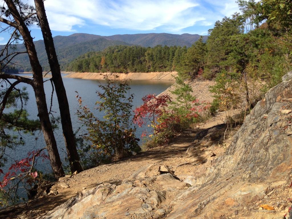 A scenic view of a lakeshore surrounded by trees and mountains, with rocky terrain leading down to the water's edge. The landscape features a mix of greenery and autumn foliage, with blue skies and wispy clouds above. Tsali Recreation Area mountain bike trail.