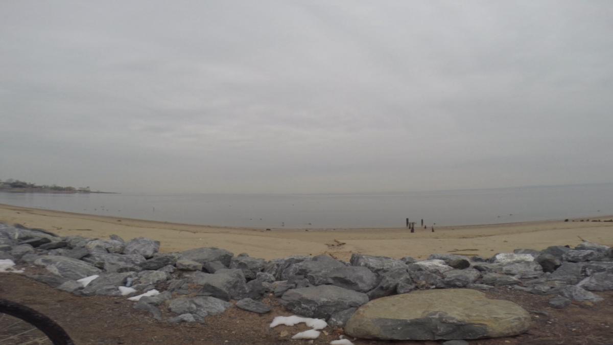 A tranquil beach scene featuring a wide stretch of sandy shoreline bordered by smooth, gray rocks. The sky is overcast with soft clouds, creating a calm and serene atmosphere. In the distance, a few wooden posts are visible along the water's edge, and small ripples can be seen on the surface of the water. The overall mood is peaceful and reflective. Wolfes Pond park mountain bike trail.