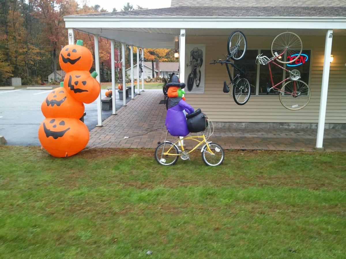 A colorful outdoor scene featuring inflatable Halloween decorations, including three stacked orange pumpkins with carved faces and a whimsical witch figure riding a yellow bicycle. In the background, two bicycles, one red and one black, are mounted on a wall near an 'Open' sign in a shop. The setting includes grass and a paved walkway under a porch, with autumn trees in the distance.
