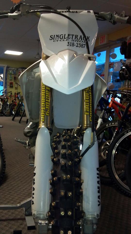 Alt text: A front view of a dirt bike displayed in a cycling shop. The bike is primarily white with gold accents on the suspension. The handlebars are positioned towards the viewer, and the tires are equipped with studs designed for traction. The shop's interior is visible in the background, containing various bicycles and cycling equipment.