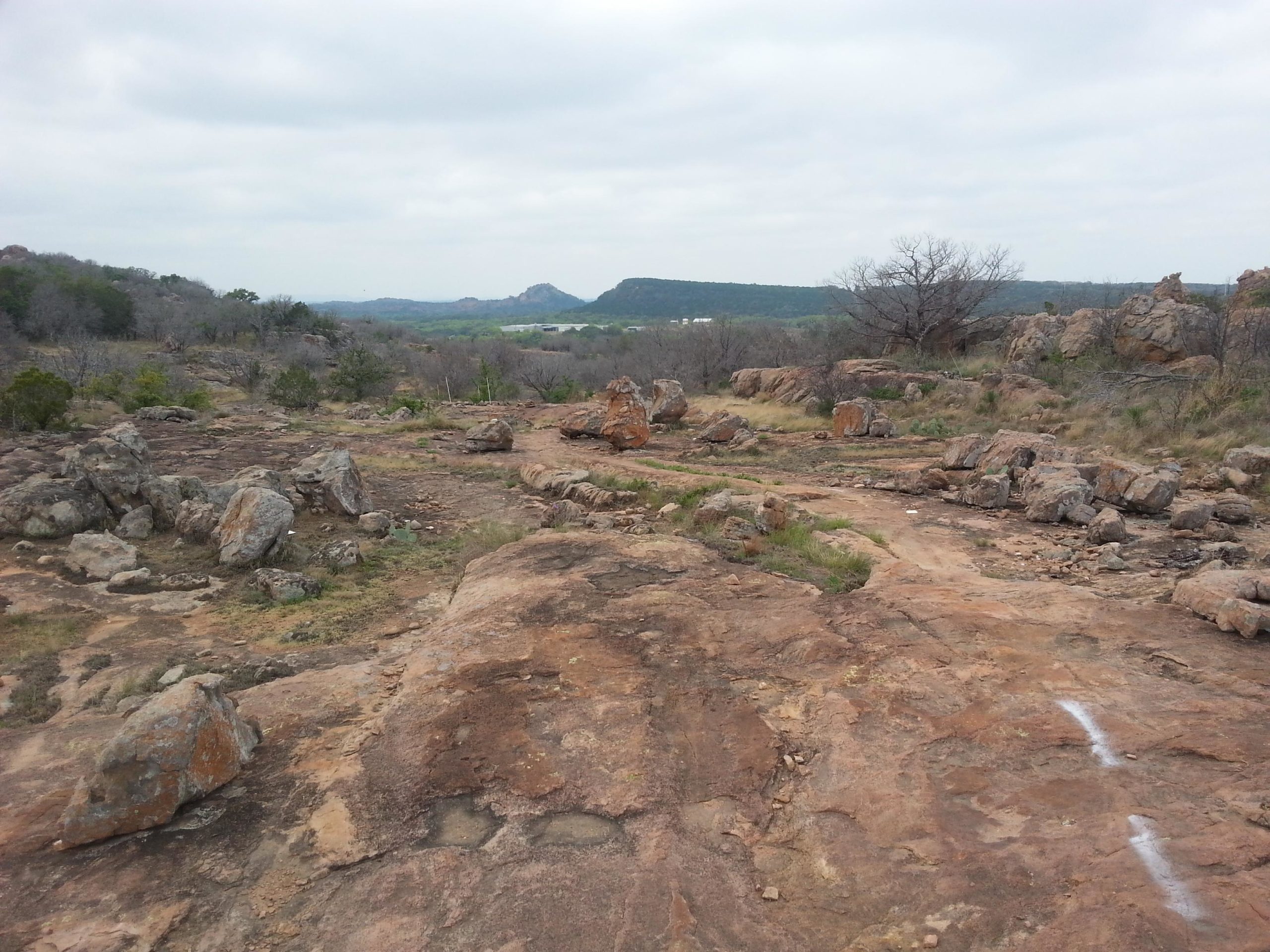 A rocky landscape featuring large boulders and sparse vegetation under a cloudy sky, with distant hills and a flat area visible in the background. The ground is predominantly rocky with patches of grass, suggesting a rugged, natural setting. Reveille Peak Ranch mountain bike trail.