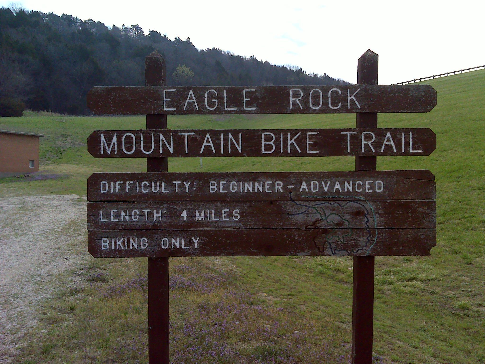 Sign for the Eagle Rock Mountain Bike Trail, indicating a length of 4 miles and difficulty level ranging from beginner to advanced. The sign specifies that the trail is for biking only and is set against a green grassy field and wooded backdrop. Eagle Rock Mountain Bike Trail mountain bike trail.