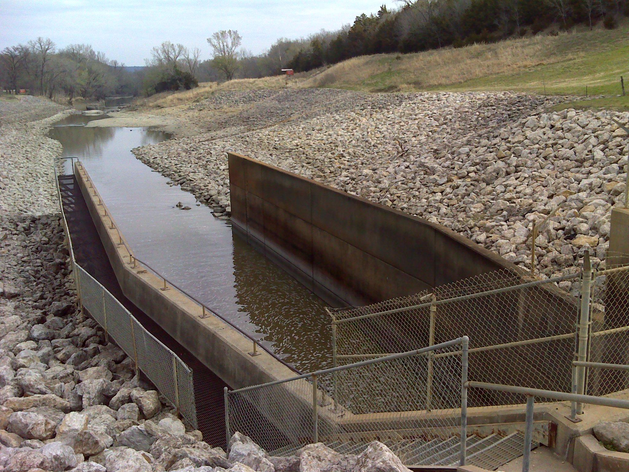 A view of a water channel surrounded by large stones, featuring a concrete structure designed for water management. The landscape includes a grassy area and scattered trees in the background, under overcast skies. A chain-link fence runs alongside the path leading to the channel. Eagle Rock Mountain Bike Trail mountain bike trail.