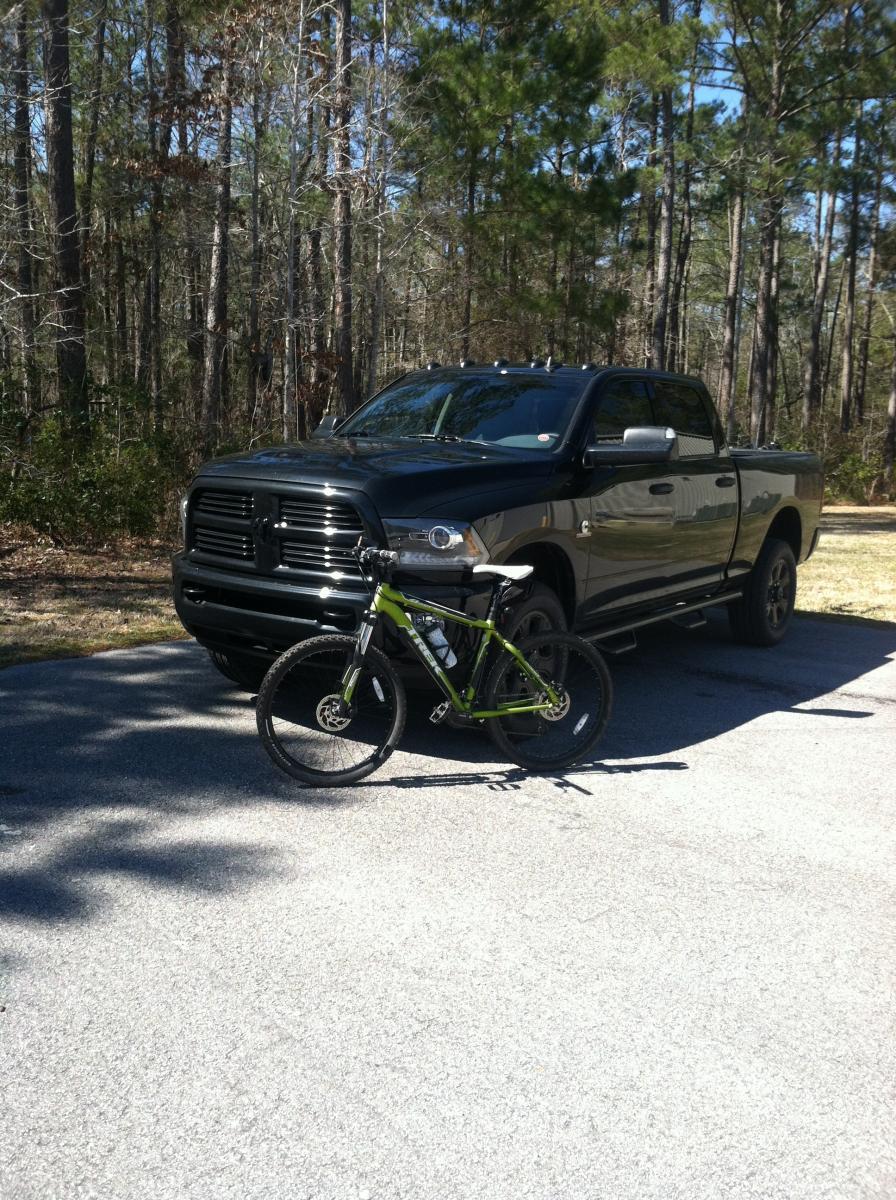 Trek 4500: A green mountain bike parked next to a black Dodge pickup truck, set against a backdrop of tall trees and a gravel road in a wooded area.