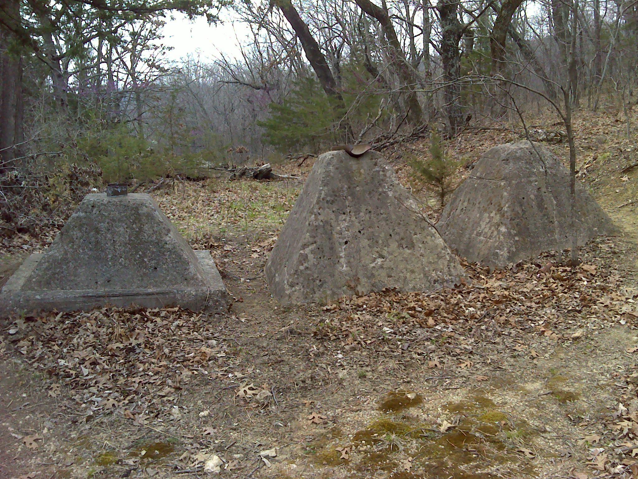 Three large, conical concrete structures are situated amidst a wooded area with scattered fallen leaves. The structures, varying in size, have a rustic appearance and are surrounded by shrubs and bare trees. The landscape is early spring, with a somewhat muted color palette of browns and greens. Eagle Rock Mountain Bike Trail mountain bike trail.