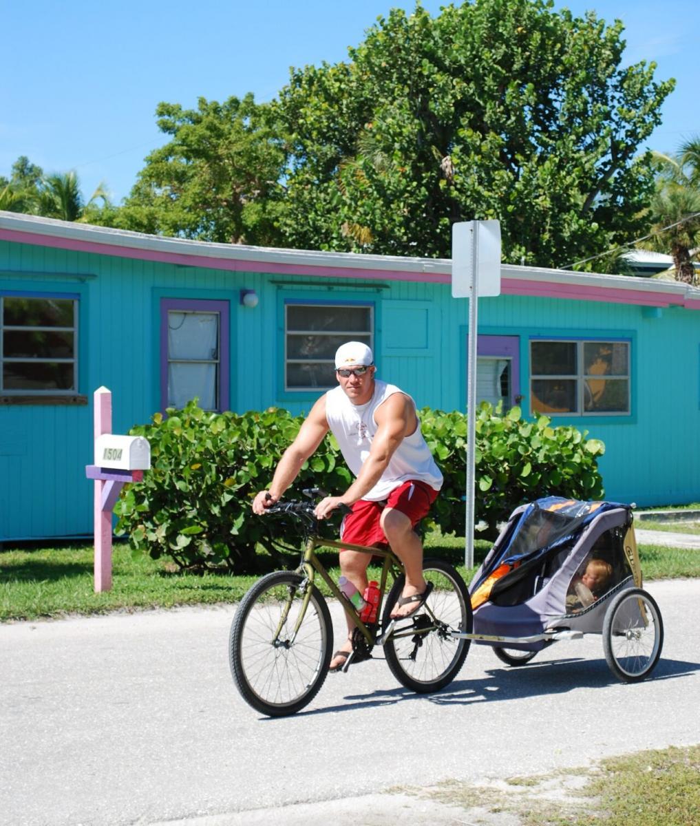 Gary Fisher Marlin: A muscular man rides a bicycle with a child trailer attached, passing by a colorful house with a blue exterior and pink accents. The scene is set on a sunny day, surrounded by lush greenery.