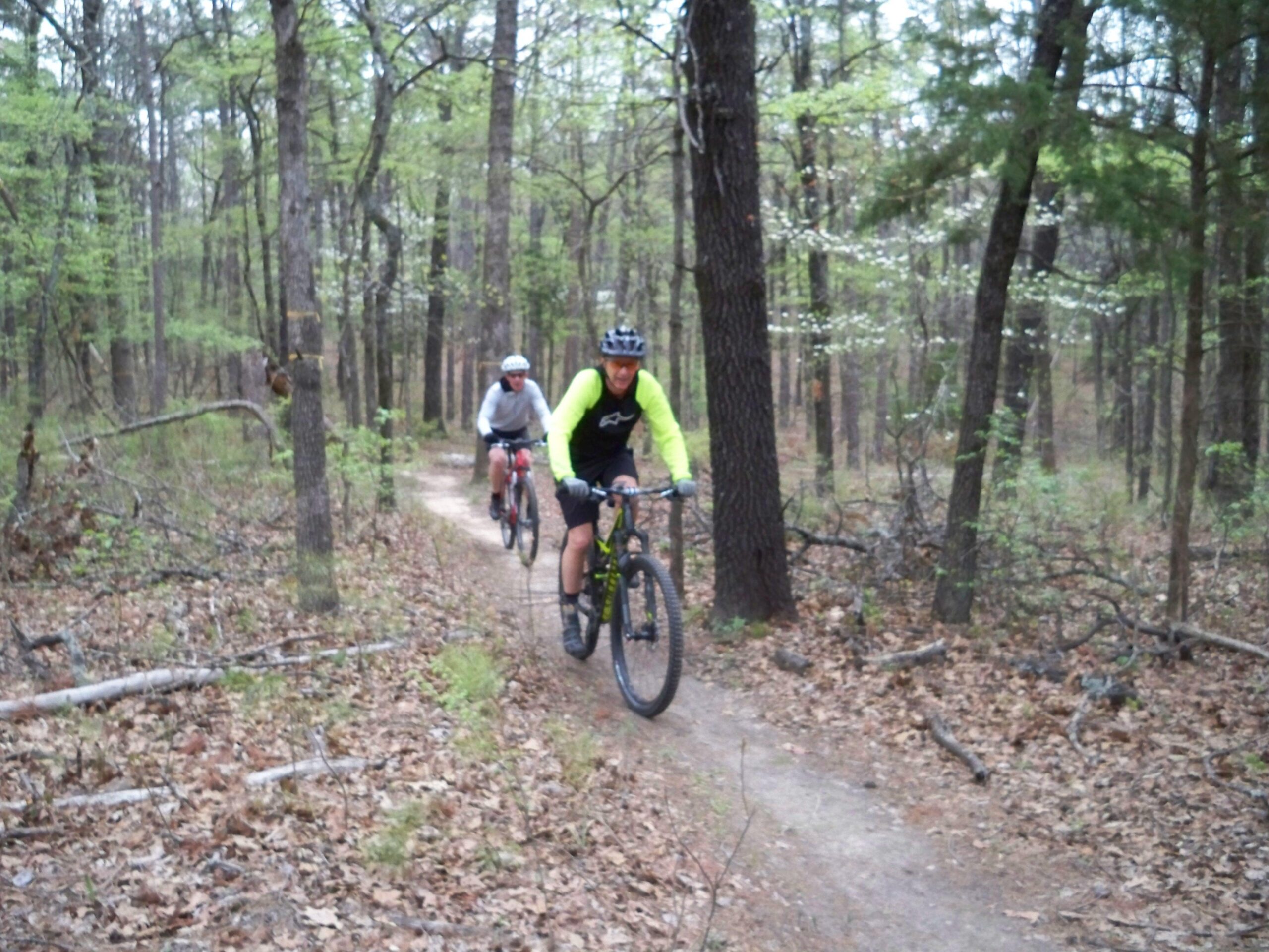 Two mountain bikers riding along a dirt trail in a wooded area. The first rider, wearing a bright green long-sleeve shirt and a helmet, is in focus, while the second rider, dressed in a white long-sleeve shirt and red shorts, follows closely behind. The scene is set among trees with fresh green leaves and scattered brown leaves on the ground, indicating an early spring atmosphere. Barber Hills Trail at Pat Mayse Lake mountain bike trail.