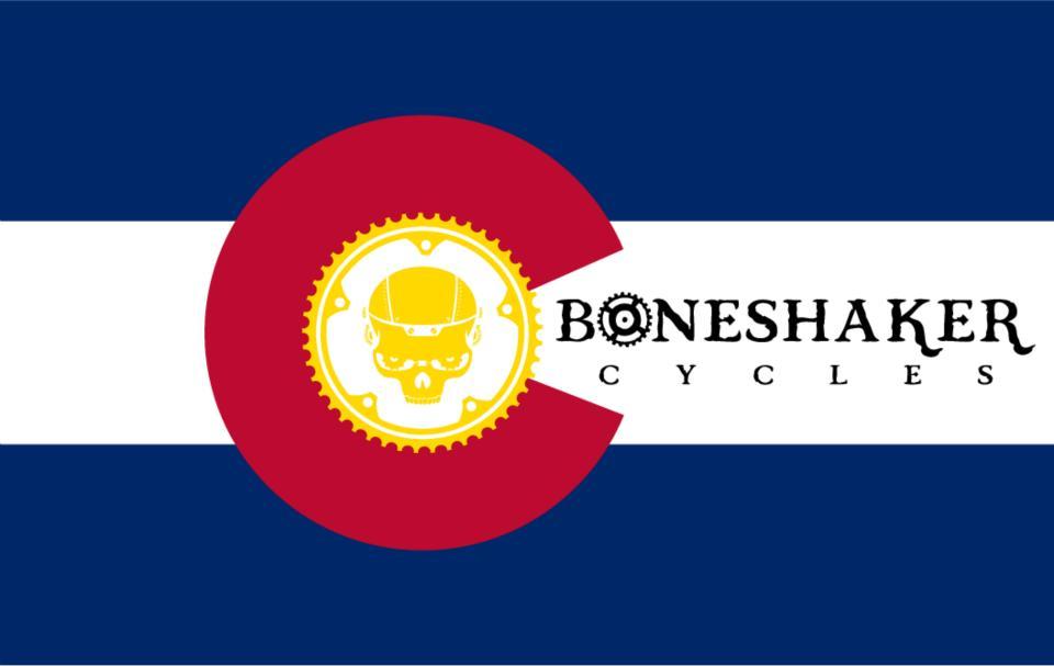 An image featuring a design inspired by the Colorado state flag, incorporating a central red circle with a yellow gear and skull icon, alongside the text "BONESHAKER CYCLES" in a vintage font. The background consists of blue and white horizontal stripes.