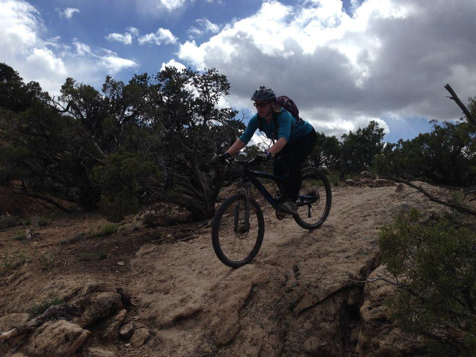 A mountain biker navigating a rocky trail, airborne as they ride over a small drop, with trees and a cloudy sky in the background. Gunny Loop mountain bike trail.