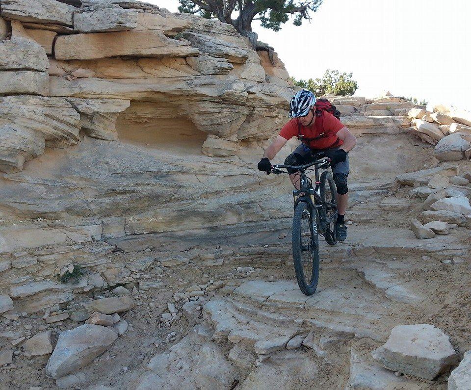 A mountain biker navigating a rocky trail with layered stone formations in a desert landscape. The rider is wearing a red shirt, gloves, knee pads, and a helmet, focused on maneuvering the bike over the uneven terrain. Mary's Loop / Horsethief Bench mountain bike trail.