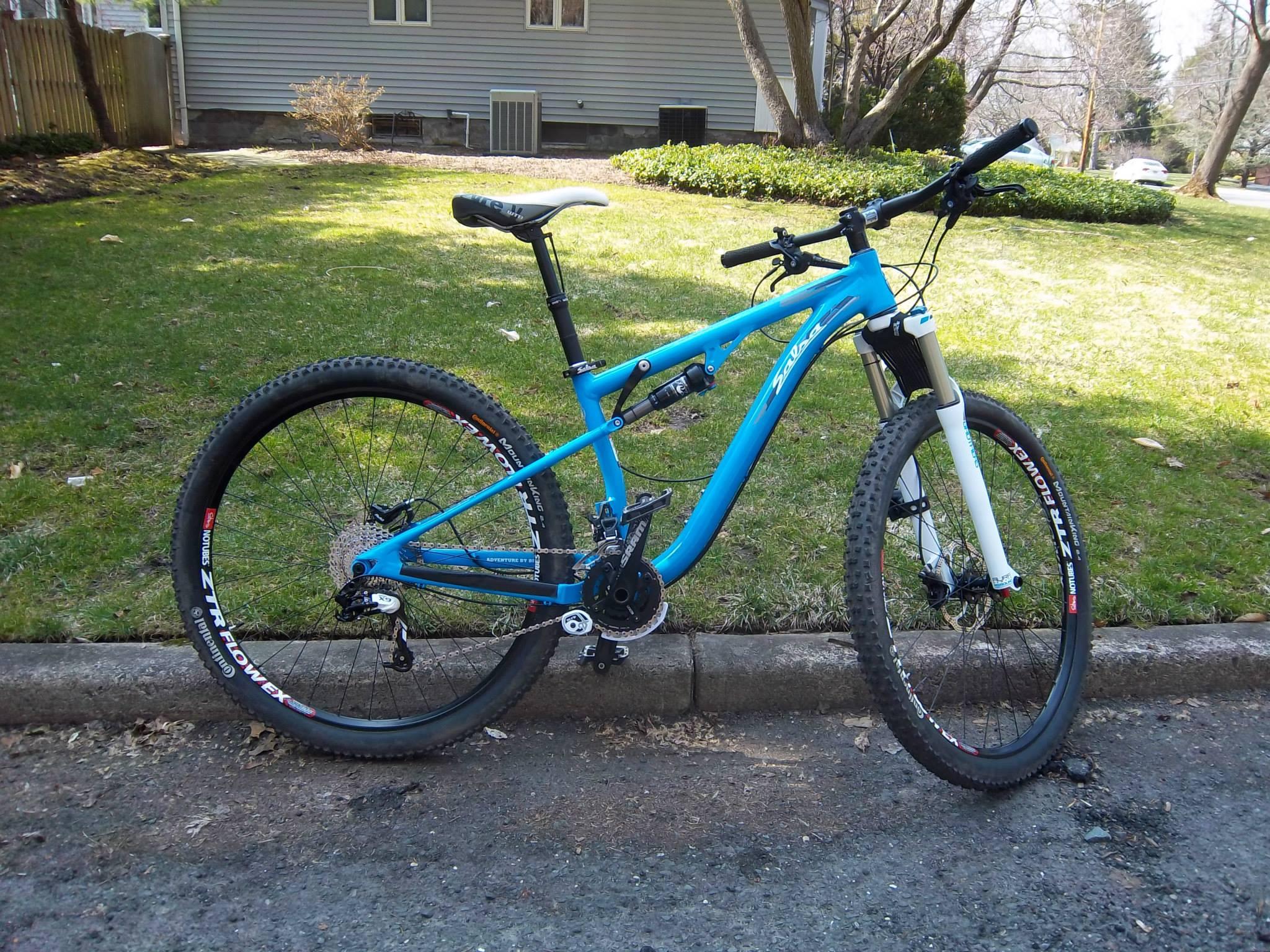 Salsa Horsethief: A blue mountain bike with a sleek frame and wide tires, parked on the side of a road. The bike features a front suspension fork and is positioned on a smooth surface, with a grassy area and a house in the background.