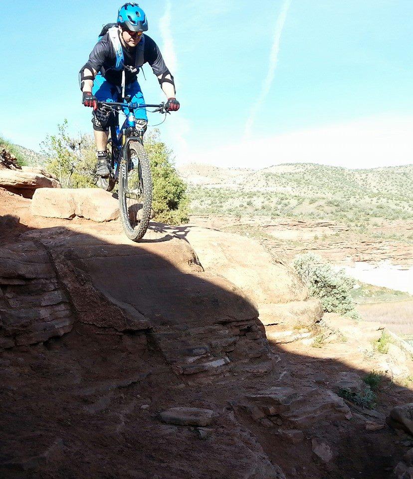 A mountain biker in blue gear jumps off a rock ledge, soaring above rugged terrain against a backdrop of hills and a clear blue sky. Mary's Loop / Horsethief Bench mountain bike trail.