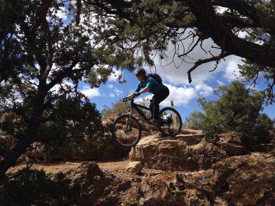 A mountain biker skillfully navigating rocky terrain under a partly cloudy sky, surrounded by trees. Gunny Loop mountain bike trail.