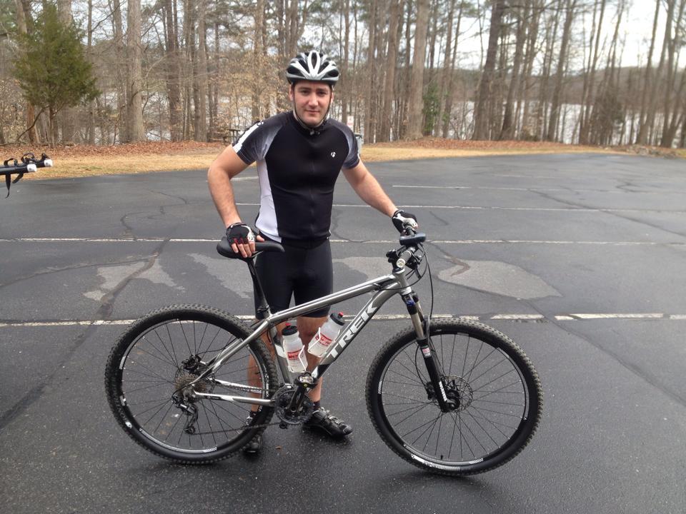 A cyclist standing next to a mountain bike in a parking area surrounded by trees, wearing a black and gray cycling outfit and a helmet. The bike has a silver frame and is equipped with two water bottles. In the background, a lake is visible through the trees. Lake Crabtree County Park mountain bike trail.