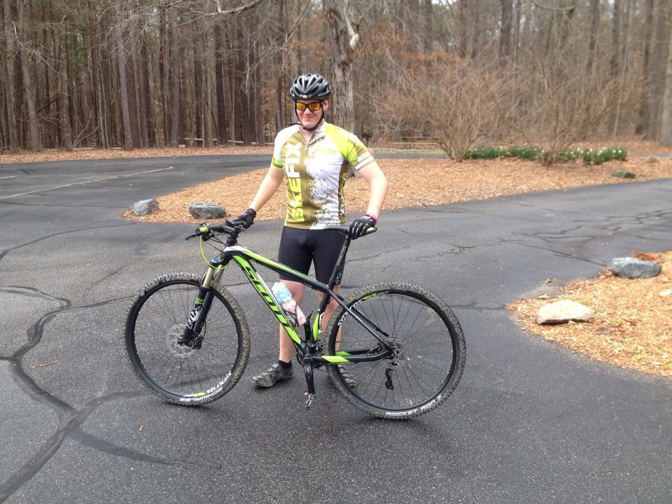 A person wearing a cycling jersey and helmet stands beside a green and black mountain bike on a paved road. The background features trees and some landscaping, with patches of bare earth and scattered rocks. Lake Crabtree County Park mountain bike trail.