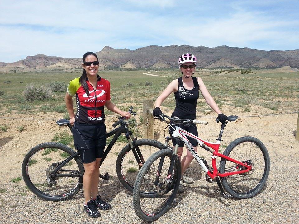 Two women stand smiling next to their mountain bikes in an outdoor setting. The background features a mountainous landscape and a clear blue sky. One woman wears a colorful cycling jersey and shorts, while the other is in a tank top with cycling gloves. They appear ready for a ride in a scenic area, surrounded by rocky terrain and open fields. Kessel Run mountain bike trail.
