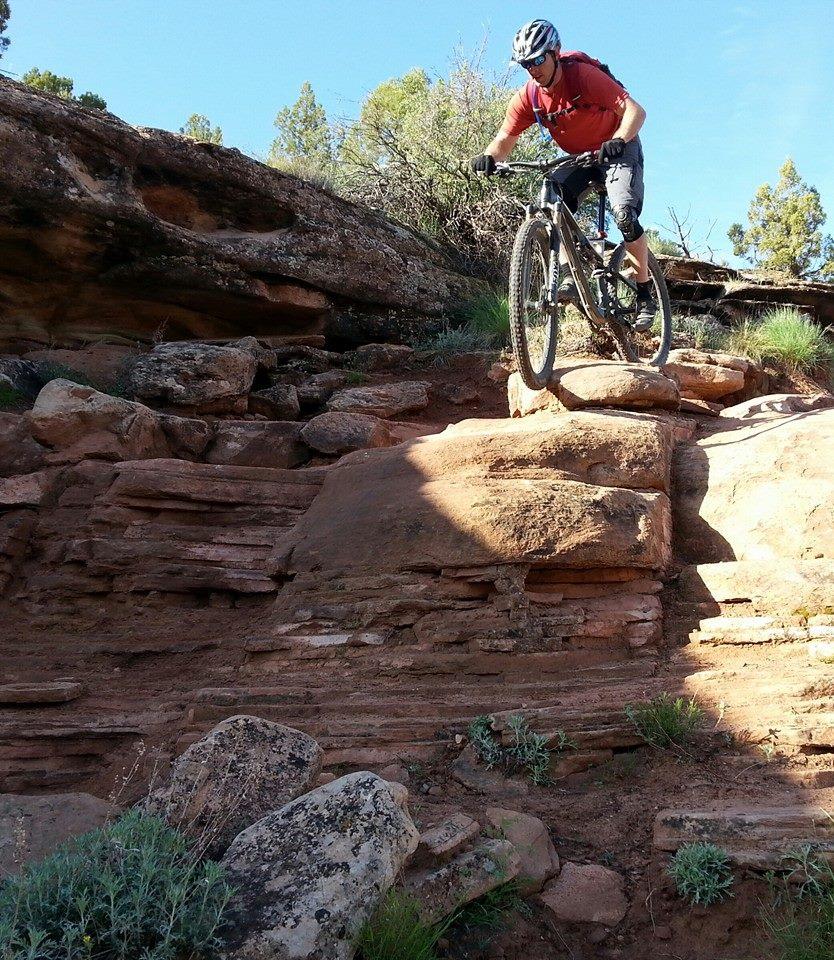 A mountain biker navigates a rocky terrain, balancing on top of a large stone while descending a steep slope. The landscape features rugged red rocks and patches of green vegetation under a clear blue sky. Mary's Loop / Horsethief Bench mountain bike trail.