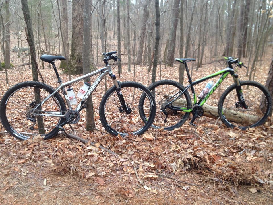 Two mountain bikes are leaning against trees in a wooded area covered with fallen leaves. The bike on the left is silver with black accents and has two water bottles mounted on the frame, while the bike on the right is predominantly green with black details. The background features tall trees and a natural forest setting. Lake Crabtree County Park mountain bike trail.