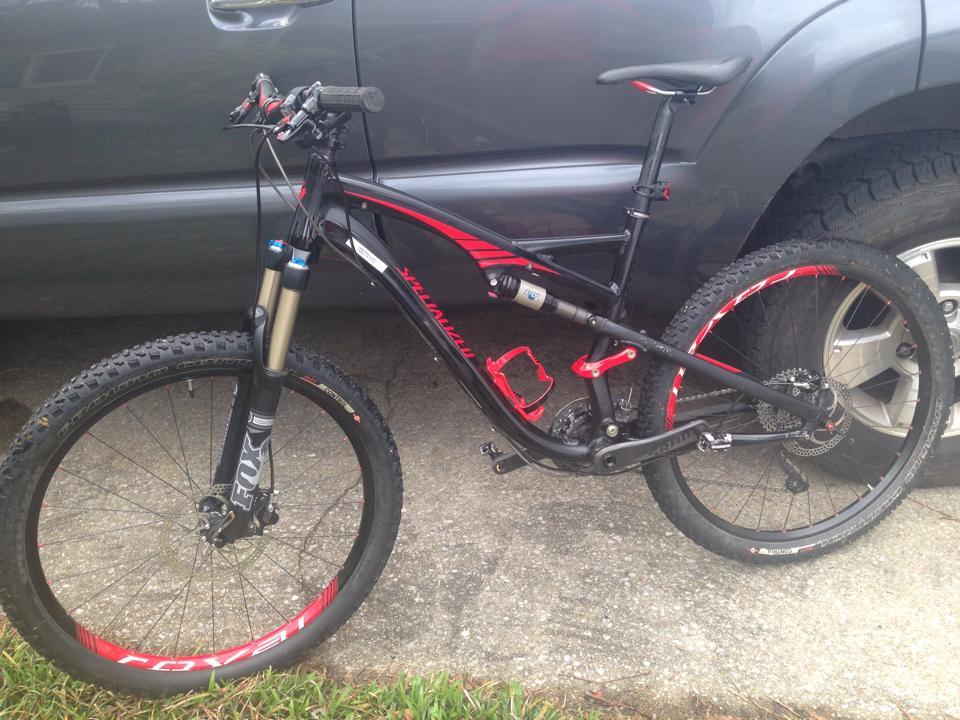 Specialized Camber Pro: A black and red mountain bike leaned against a gray vehicle, featuring thick tires and a front suspension fork. The bike is equipped with a bottle cage and has a modern design suitable for off-road riding. The background shows a concrete surface with grass on the side.