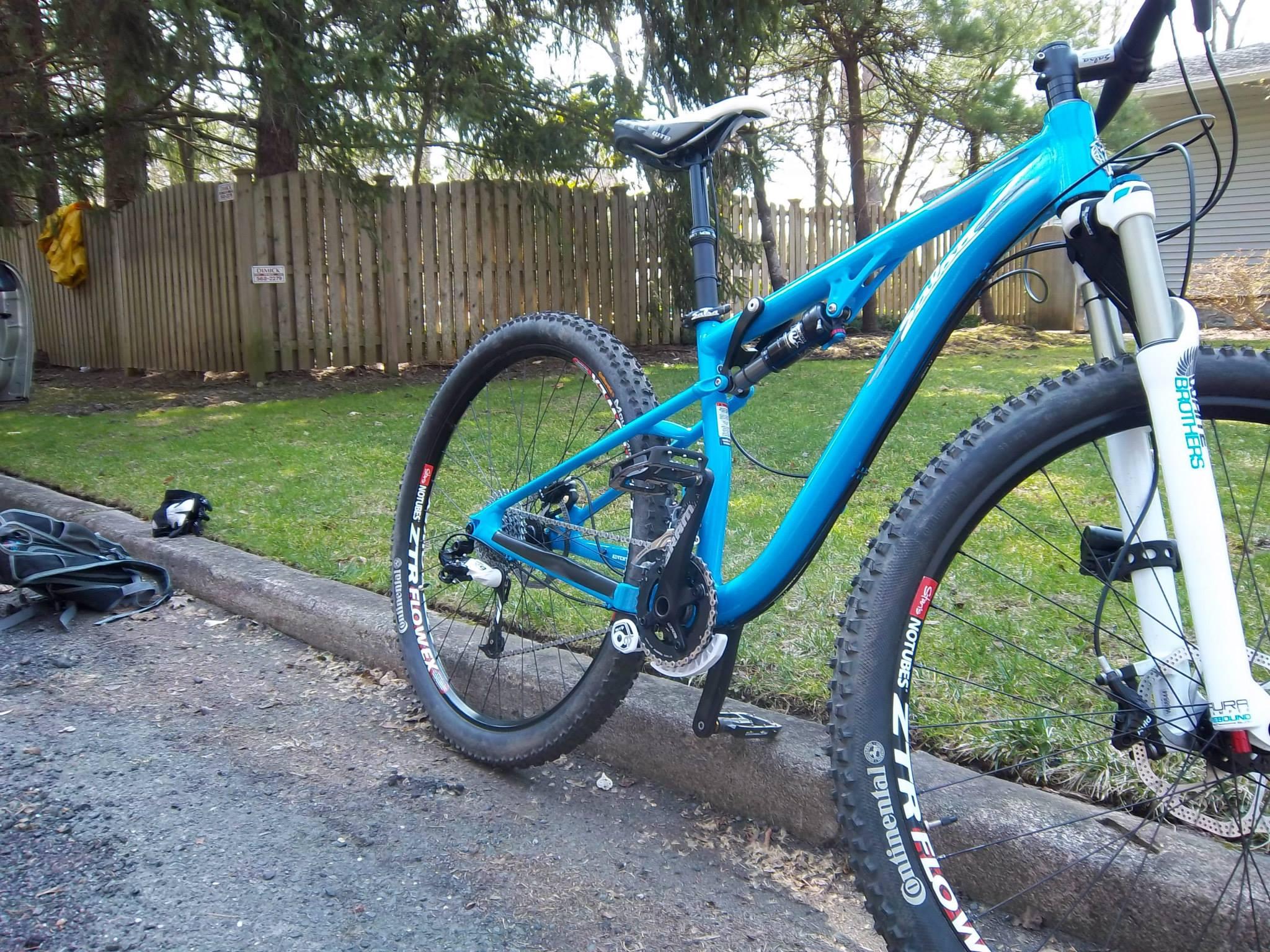Salsa Horsethief: A blue mountain bike with thick tires parked near a grassy area, showing the bike's front suspension and gear mechanisms. In the background, there is a wooden fence and some scattered outdoor gear.