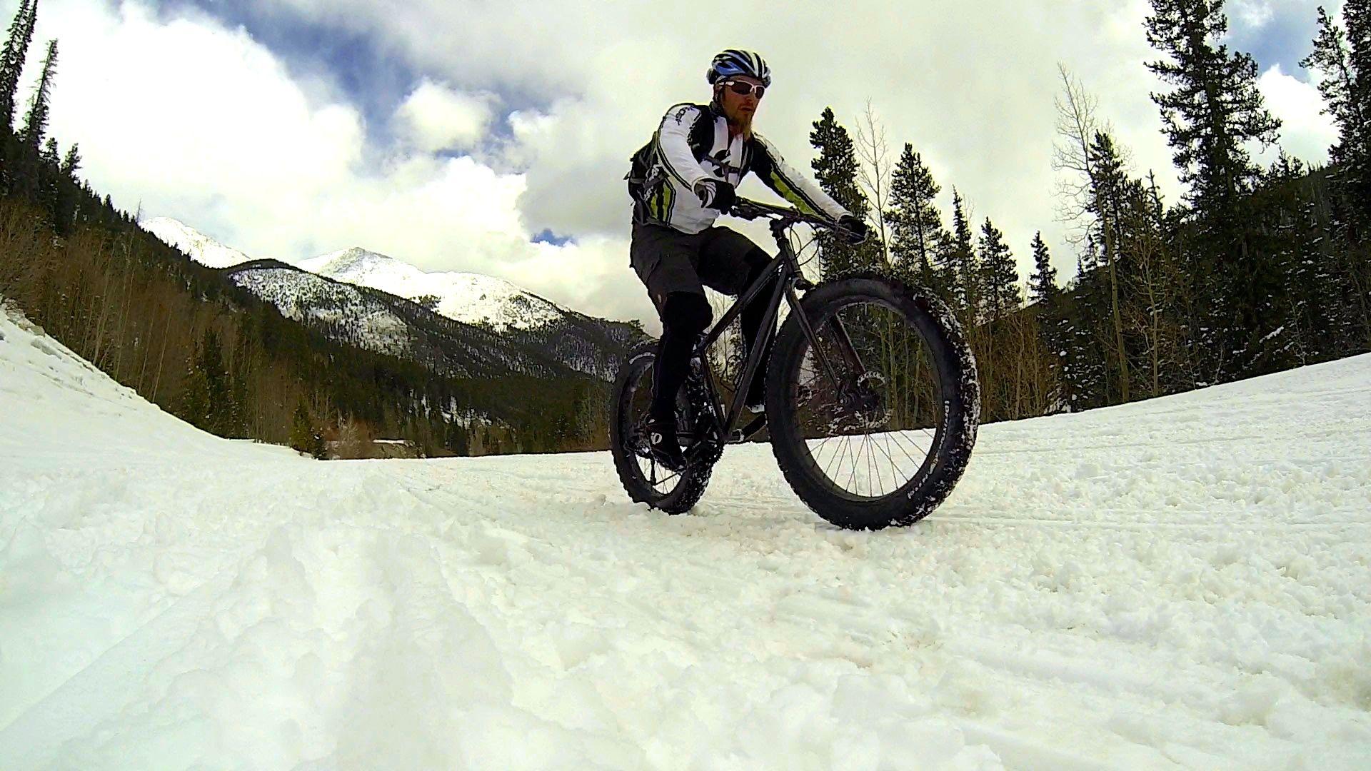 A person riding a fat-tire bicycle on a snow-covered path with mountains in the background. Pine trees line the trail under a partly cloudy sky. The cyclist is wearing winter gear, including gloves and sunglasses. Cottonwood Pass Road mountain bike trail.
