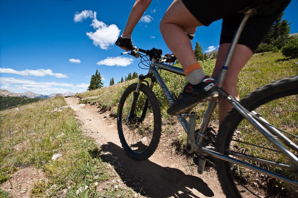 A cyclist riding a mountain bike along a dirt trail surrounded by greenery and rocky terrain under a clear blue sky with scattered clouds. Monarch Crest Trail mountain bike trail.