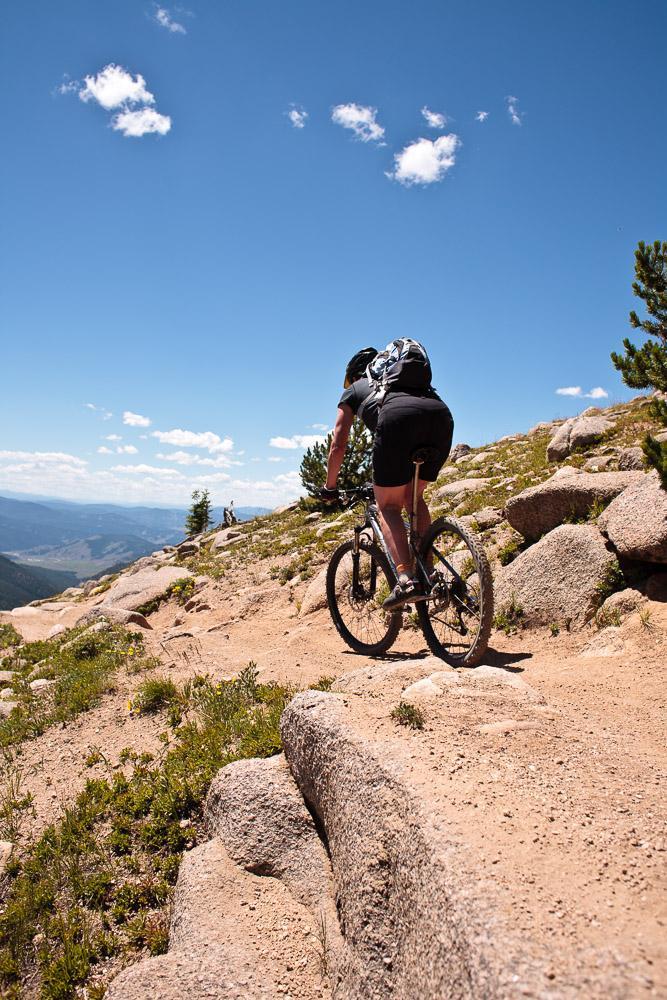 A mountain biker navigating a rocky trail under a clear blue sky, with green vegetation and distant mountains visible in the background. Monarch Crest Trail mountain bike trail.