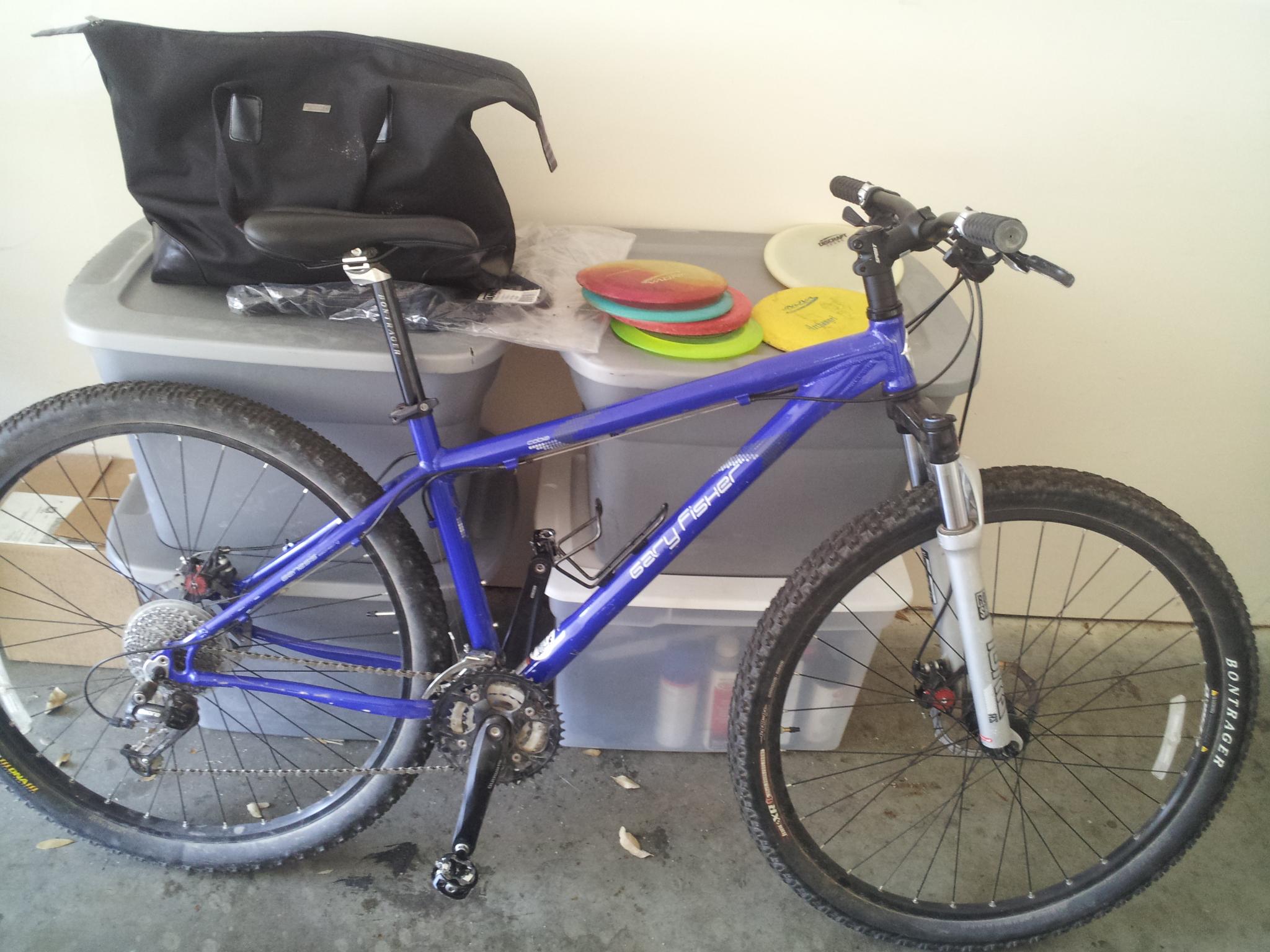 Gary Fisher Cobia: Mountain bike in a garage setting, featuring a blue frame, black tires, and disc brakes. In the background, there are plastic storage bins and colorful frisbees. A black bag is also placed on top of one of the bins.