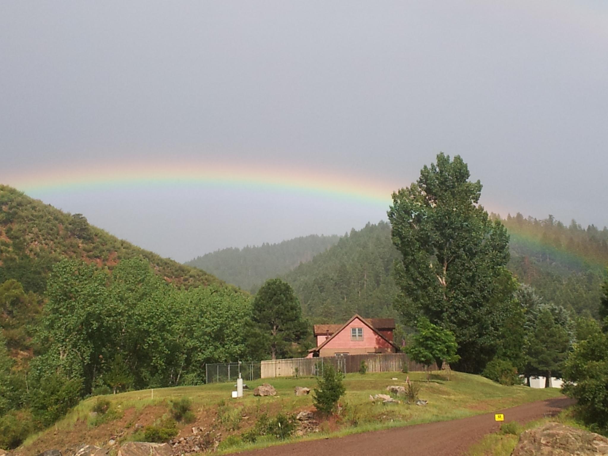 A vibrant rainbow arches over a mountainous landscape, with a pink house nestled among green trees. The scene features rolling hills covered in lush vegetation, under a partly cloudy sky. A gravel road leads toward the house, enhancing the tranquil rural setting. Colorado Trail: Waterton To South Platte mountain bike trail.