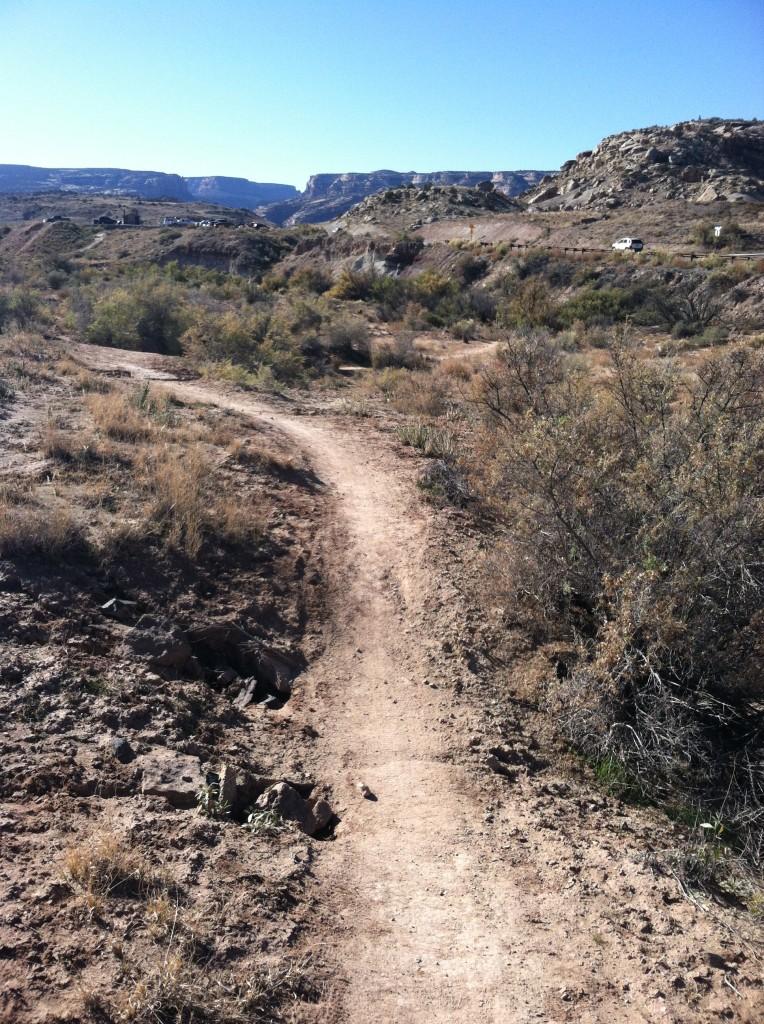 A dirt trail winding through a dry, natural landscape with sparse vegetation and rocky terrain, leading towards a distant view of cliffs and hills under a clear blue sky. A vehicle is visible along a road to the right. Yes N DeeDee mountain bike trail.
