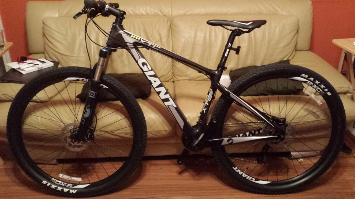 Giant XTC 29er: A black and white mountain bike is leaning against a beige couch. The bike features thick tires with "Maxxis" branding, a front suspension fork, and a sleek design with prominent "GIANT" and "XTC" logos. A brown pillow is visible on the couch, along with various items on a nearby table. The image has warm indoor lighting.