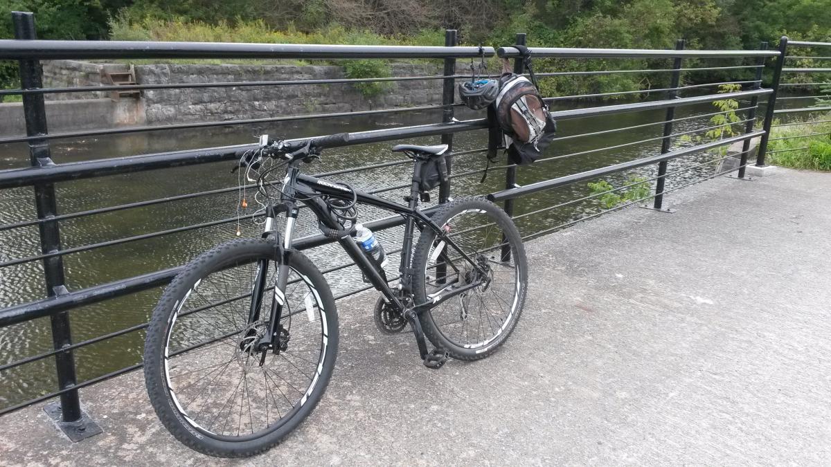 Specialized Rockhopper Comp 29: A black mountain bike with a water bottle is parked on a concrete path near a railing that overlooks a calm body of water. A small backpack hangs from the railing, and greenery can be seen in the background.