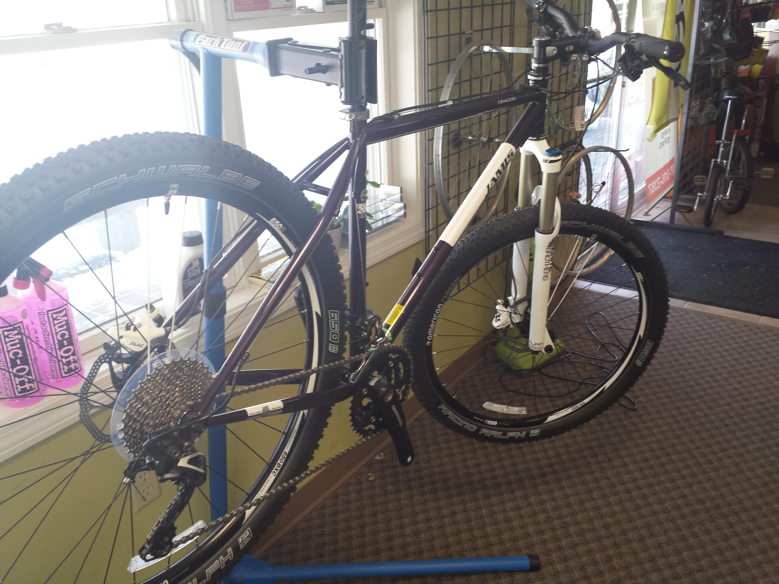 A purple and white mountain bike is displayed in a bike shop. It is mounted on a repair stand, showcasing its detailed components, including the wheels with Schwalbe tires and a rear cassette. The background features a shop interior with bike accessories and parts visible.