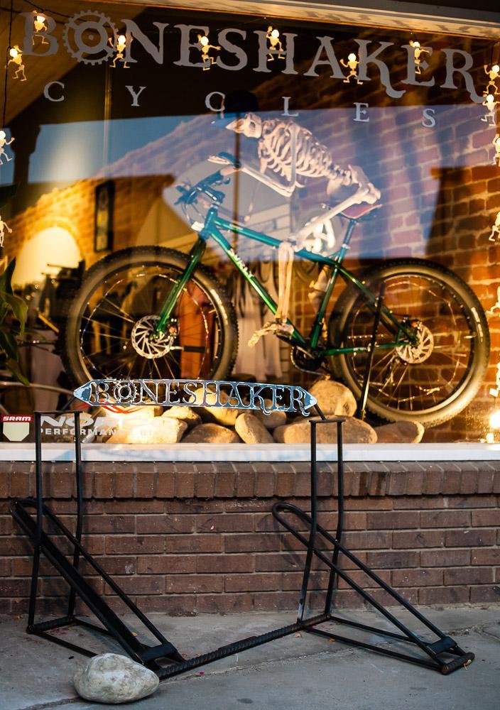 Alt text: A storefront for Boneshaker Cycles featuring a large sign and a green bicycle displayed in the window. In front of the store, there is a black bike rack with a decorative "Boneshaker" sign, positioned on a brick sidewalk. The scene is illuminated by string lights.
