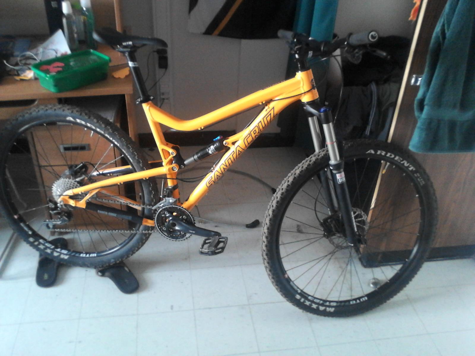 Santa Cruz Tallboy LT: A bright orange mountain bike is leaning against a desk in a room, showcasing a sleek frame and rugged tires. The bike features a suspension fork, multiple gears, and wide tires suitable for off-road cycling. In the background, there are a few personal items on the desk and a closet with hanging clothes.