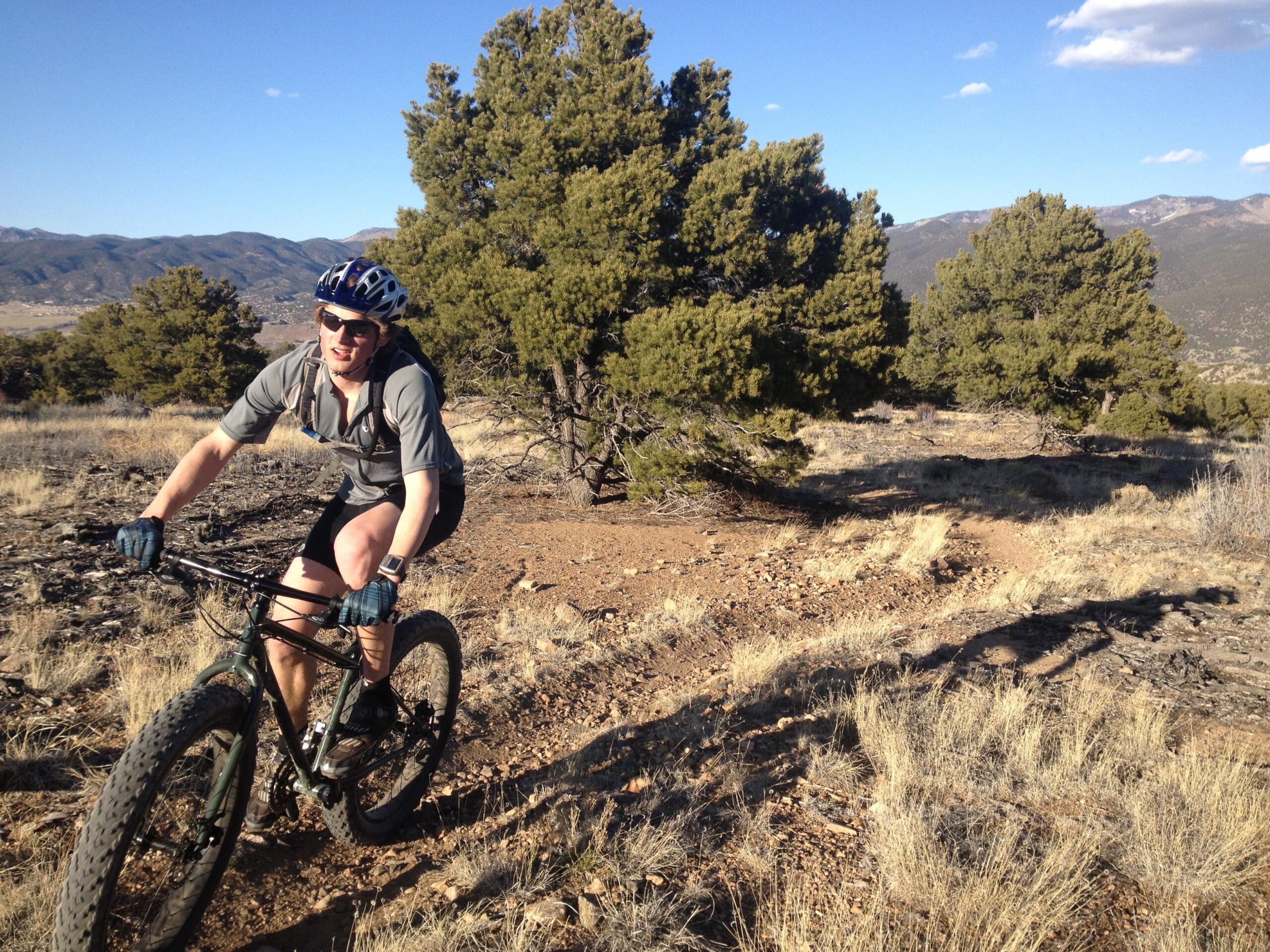 A person riding a mountain bike on a dirt trail surrounded by tall trees and mountainous terrain. The cyclist, wearing a helmet and sunglasses, appears to be enjoying the ride in a sunny outdoor setting. Methodist Mountain mountain bike trail.