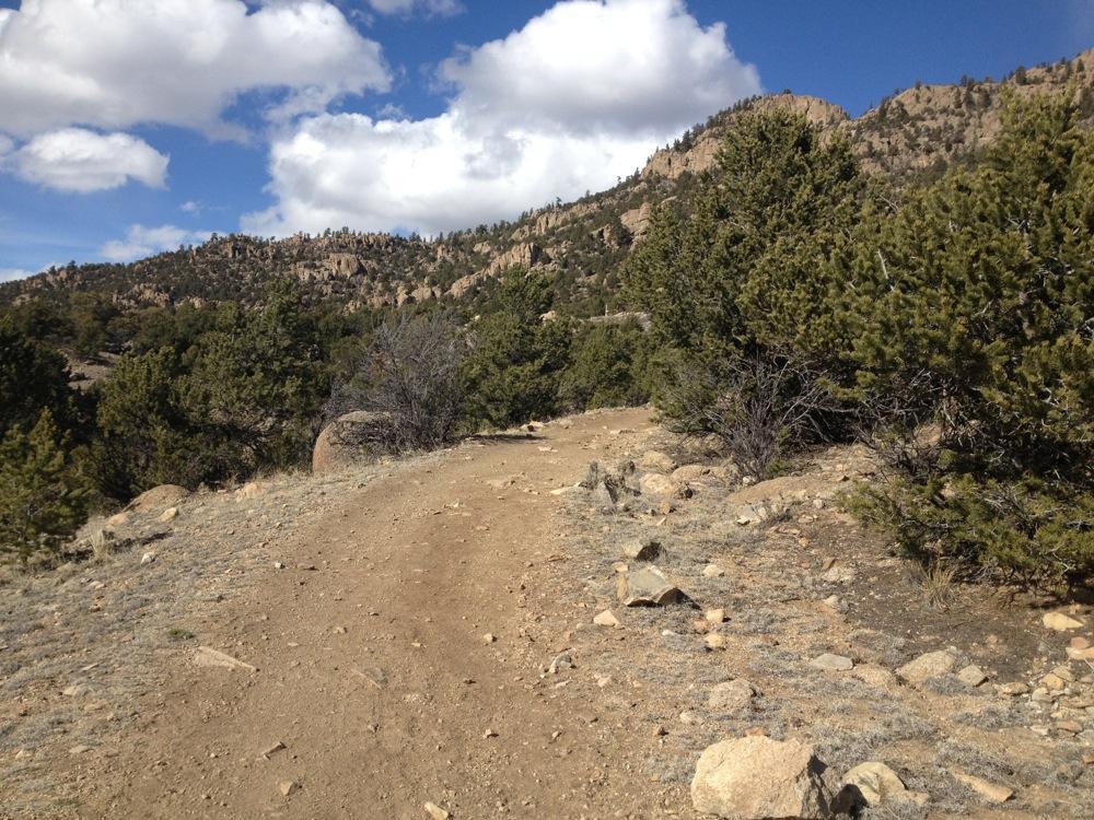 A dirt trail winding through a rocky landscape surrounded by pine trees, with hills and a partly cloudy sky in the background. Whipple Trail mountain bike trail.