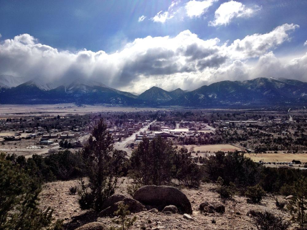 A panoramic view of a mountainous landscape under a partly cloudy sky. In the foreground, scattered rocks and shrubs are visible, leading to a small town in the valley below. The town is surrounded by open fields and dotted with buildings, with majestic mountains rising in the background. Snow can be seen on the mountain peaks, indicating a winter setting. Whipple Trail System mountain bike trail.