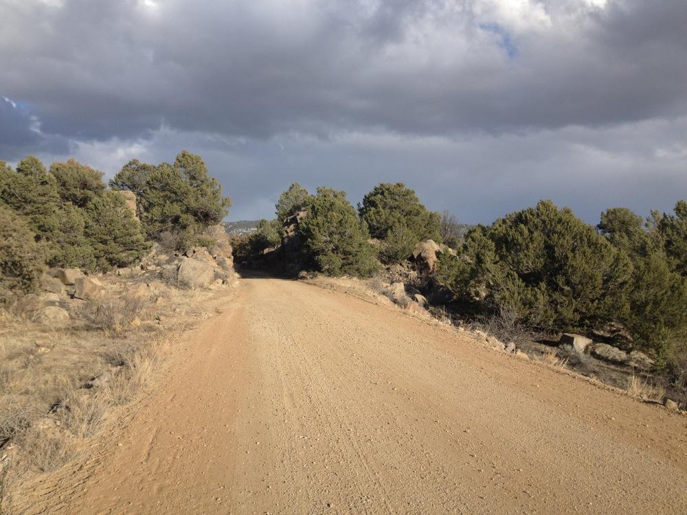 A dirt road winding through a landscape of sparse vegetation and rocky outcrops, with a cloudy sky overhead. The scene captures the natural beauty of a rugged outdoor setting, ideal for adventures or exploration. Midland Hills Trails mountain bike trail.