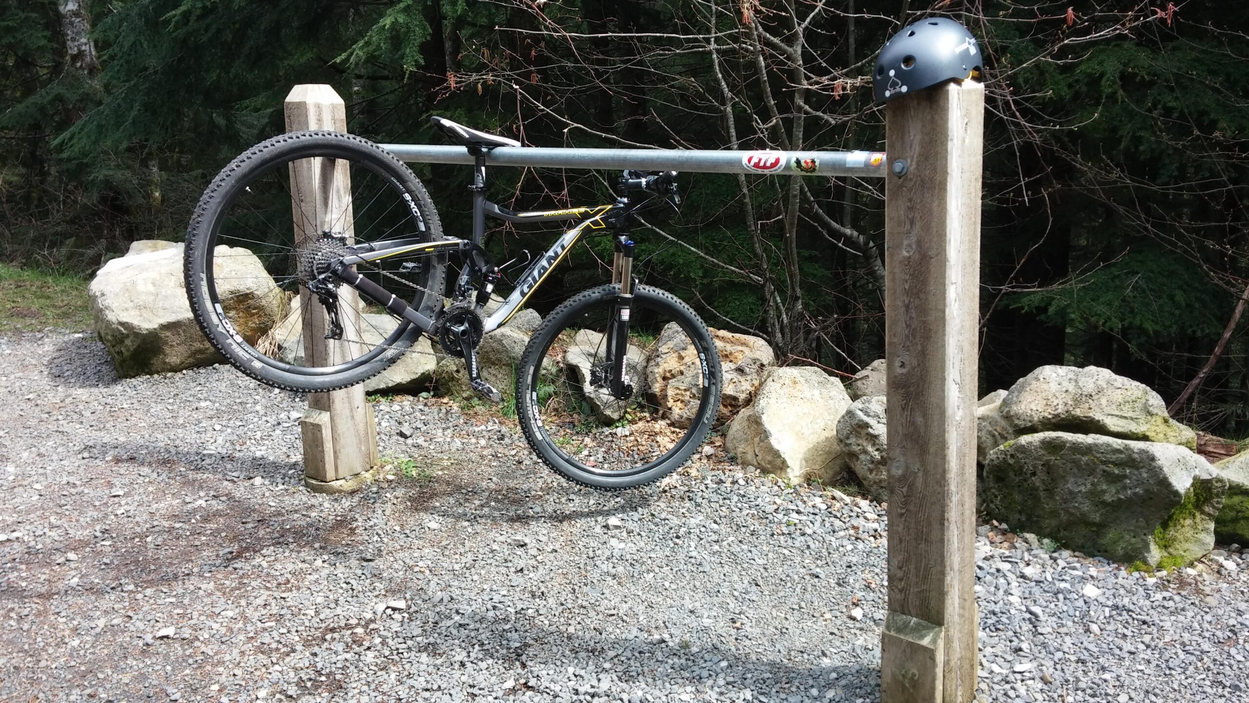 Giant Trance X 29er 1: A mountain bike hanging on a horizontal railing at a trailhead, with a black helmet resting on a nearby wooden post. The ground is gravel with scattered rocks, and green trees are in the background.