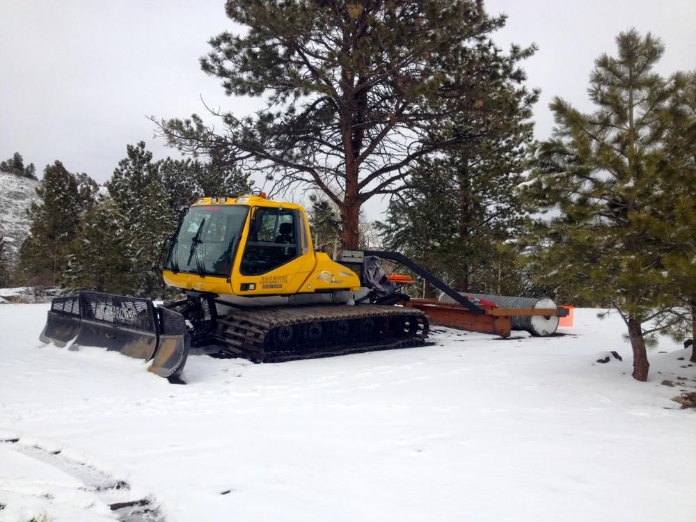 A yellow snowcat vehicle parked on a snowy landscape, surrounded by pine trees. The snowcat has a large blade attached to the front for snow removal and a roller at the rear. The scene is set in a winter environment with a clear sky and snow-covered ground. Marshall Pass Road / #200 / #203 / #243 mountain bike trail.