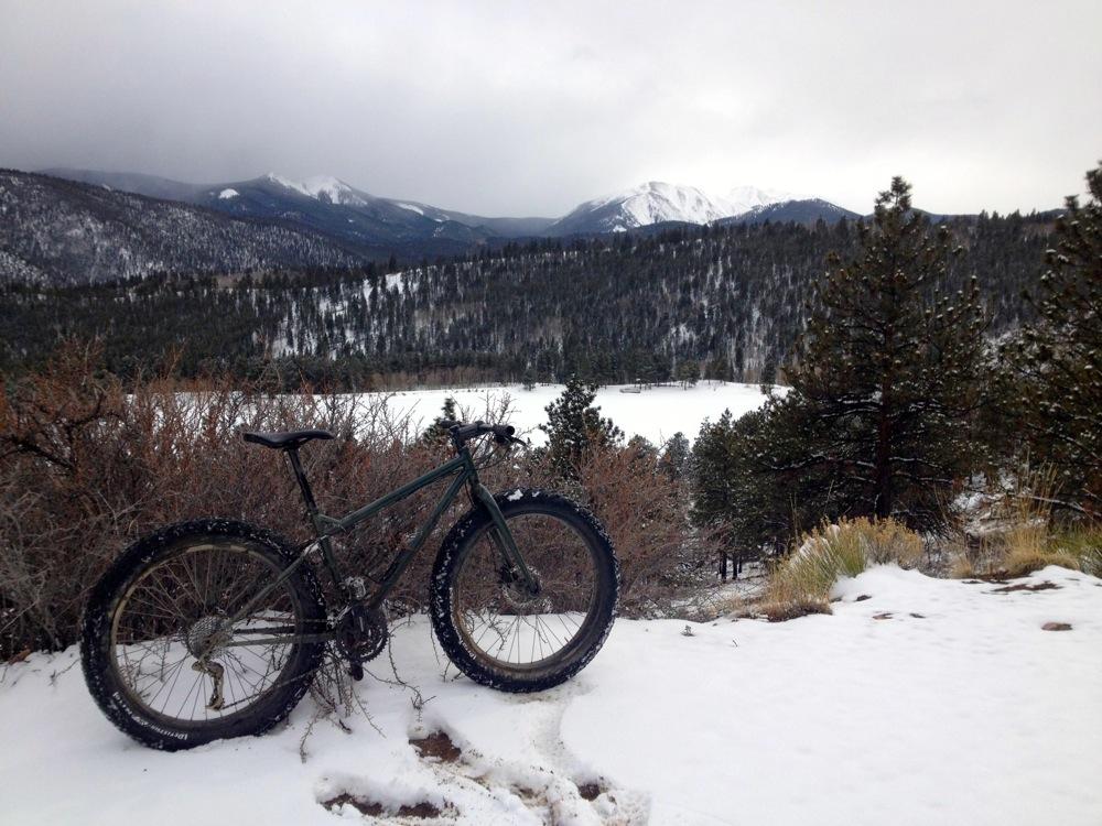 A mountain bike rests on a snowy trail overlooking a scenic landscape of snow-covered mountains and a forested area with a partially frozen lake. The sky is overcast, creating a chilly winter atmosphere. Marshall Pass Road / #200 / #203 / #243 mountain bike trail.