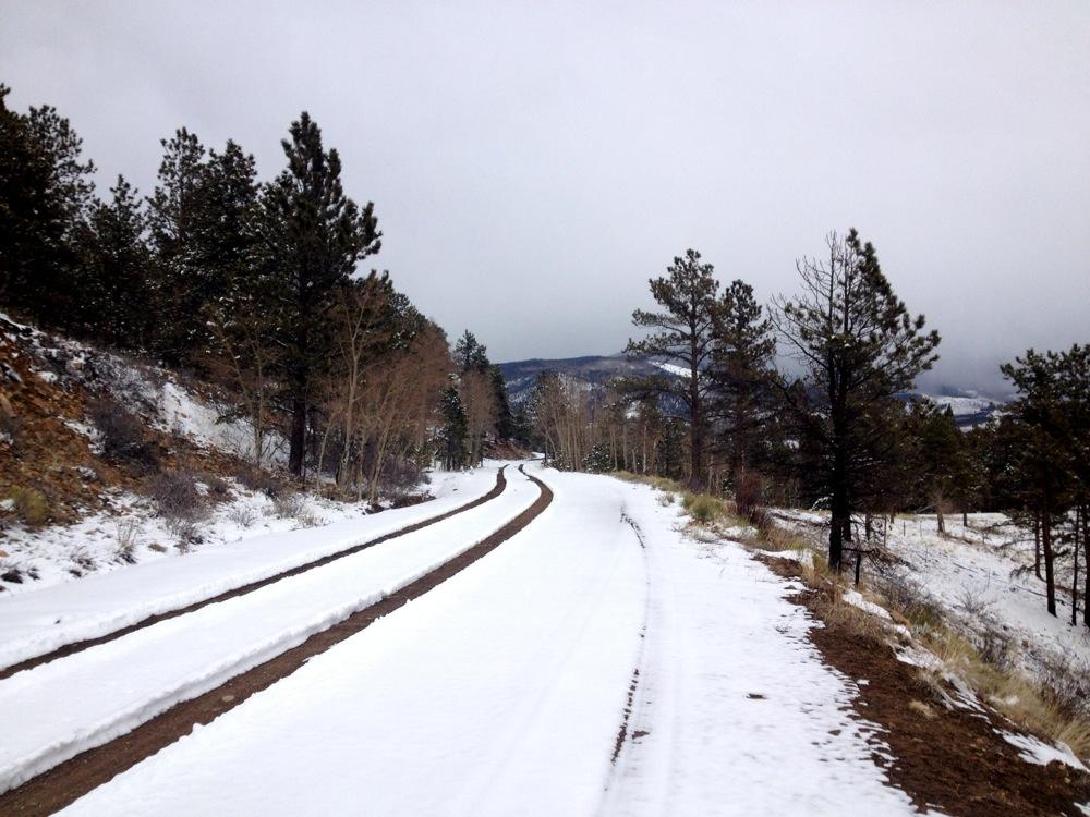 A snow-covered dirt road winding through a mountainous landscape, flanked by evergreen trees and aspen groves, under a cloudy sky. Marshall Pass Road / #200 / #203 / #243 mountain bike trail.