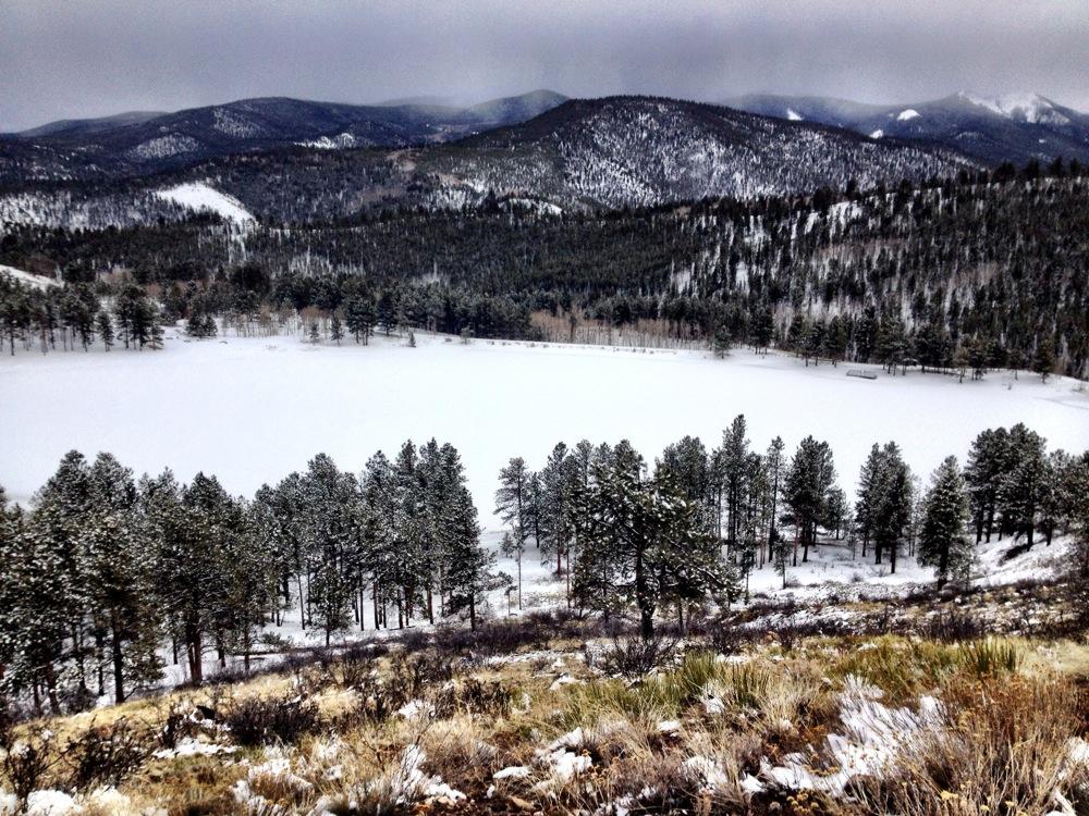 A snowy landscape featuring a frozen lake surrounded by evergreen trees, with distant mountains and a cloudy sky in the background. The scene captures the serene beauty of winter in a forested area. Marshall Pass Road / #200 / #203 / #243 mountain bike trail.