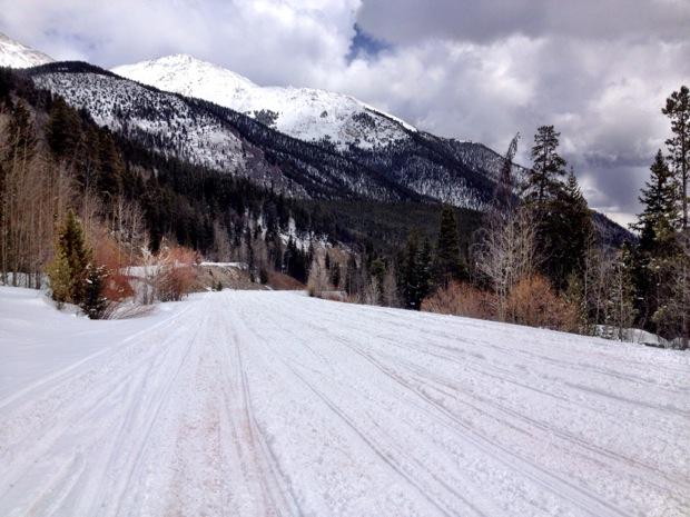 Snow-covered landscape with a wide path running through it, framed by evergreen trees and mountains in the background. The sky is partly cloudy, adding a serene atmosphere to the winter scene. Cottonwood Pass Road mountain bike trail.